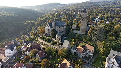 Luftaufnahme der Burg Kronberg. Sie liegt oberhalb von Kronberg im Taunus und ist eingebettet in viel Wald. Der Bergfried befindet sich etwas separat von den weiteren Burgteilen auf der rechten Seite. Unterhalb von der Burg ist außerdem noch der Rosengarten zu sehen. Die Burg ist sehr gut erhalten. 