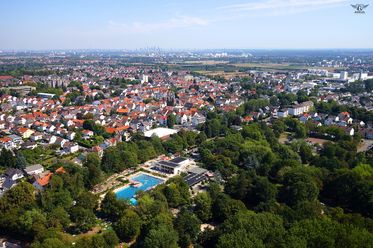 Das Foto ist eine Luftaufnahme von Kriftel. Im Vordergrund ist das Parkbad zu sehen, das sich wie der Name schon sagt in einem Park mit zahlreichen Bäumen befindet. Auf der hinteren linken Seite des Fotos ist Kriftel zu sehen. Am Horizont sieht man ganz klein noch die Skyline von Frankfurt. 