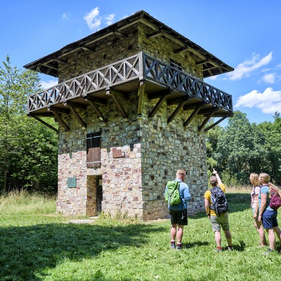 Eine Wandergruppe steht vor dem vollständig restaurierten Wachturm des Kastell Zugmantel. Der Himmel ist blau, im Hintergrund stehen grüne Böume, die Wanderer stehen auf einer Wiese vor dem Wachturm.