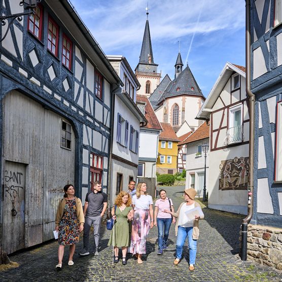 Das Foto zeigt eine kleine Gruppe, die gerade eine Stadtführung macht. Sie befinden sich in einer Gasse mit vielen Fachwerkhäusern. Im Hintergrund ist die St. Ursula Kirche zu sehen. 
