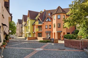 Das Foto zeigt die Außenfassade der Bücherei in Hattersheim. Der Straßenbelag ist hauptsächlich aus Kopfsteinpflaster. Der Bereich ist eine Fußgängerzone und hat hier und da Treppenstufen. 