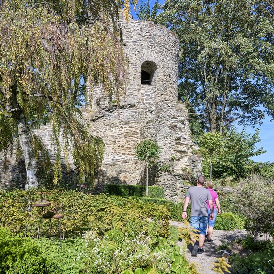 Burg Altweilnau ist noch als Ruine erhalten. Das Paar auf dem Foto ist auf dem Weg dorthin und geht durch dicht gewachsene Büsche und Bäume auf einem schmalen Pfad. Der Himmel ist strahlend blau an einem sonnigen Sommertag. 