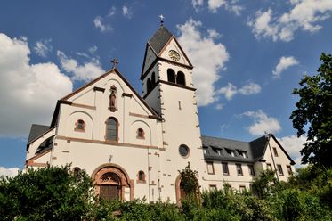 Auf dem Foto ist die Frontansicht der Kirche St. Franziskus in Kelkheim zu sehen. Das Foto wurde von unten aufgenommen. Die Fassade der imposanten Kirche ist weiß mit Details aus rotem (Sand)stein. Neben dem Kirchenschiff befindet sich rechts der Kirchturm. 