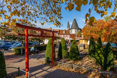 Der Platz am Untertor in Hofheim befindet sich in der Altstadt. Aus roten Balken ist das Tor nachgestellt. Im Hintergrund ist ein kleines Türmchen zu sehen. Die Aufnahme wurde im Herbst gemacht, die Blätter der Bäume rund herum sind schon bunt gefärbt. 