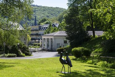 Im Vordergrund sind zwei Storchskulpturen zu sehen, die auf einer Wiese stehen. Im Hintergrund befinden sich mehrere schmucke Gebäude. 