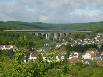 Eingebettet in zahlreiche Bäume liegt in einer Talsenke Niedernhausen. Hinter dem Taunusort sieht man die Theißtalbrücke, eine große Autobahnbrücke der A3