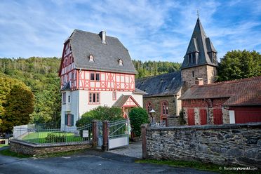 Das Foto zeigt das Pfarrhaus in Rod an der Weil, das im oberen Bereich noch rotes Fachwerk hat, im unteren Bereich ist es weiß verputzt. Direkt daneben steht die kleine Kirche aus braunen Steinen. Außen herum ist eine halbhohe Mauer aus Steinen zu sehen. 