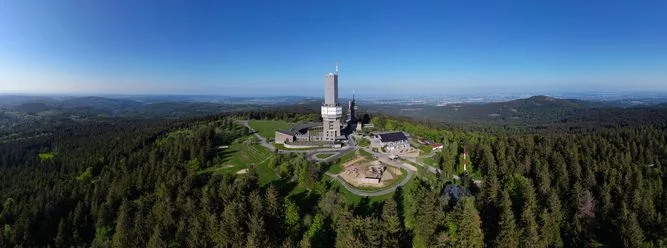 Panoramaaufnahme des Großen Feldberg. Zu sehen sind auch alle Gebäude, die auf dem Plateau stehen, wie Funkturm des hr und der Aussichtsturm. Außen herum ist die hügelige Waldlandschaft des Taunus zu sehen. Der Himmel ist strahlend blau. 