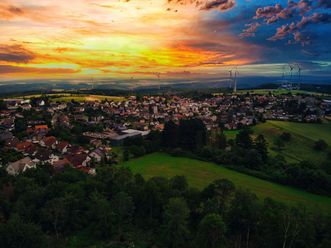 Das Foto ist recht dunkel, im Vordergrund ist Natur zu sehen mit Feldern und Wald, im linken hinteren Bereich der kleine Ort Heidenrod. Im Hintergrund ist der Himmel vom Sonnenuntergang gelb-orange gefärbt. 