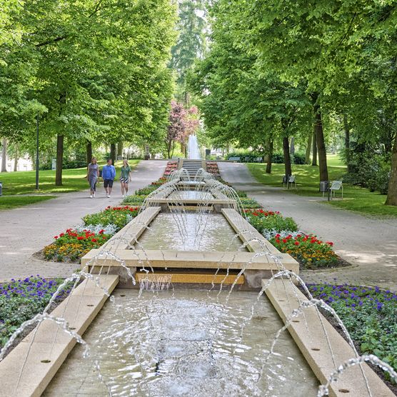 der langgezogener Brunnen mit meheren Ebenen befindet sich im Kurpark von Bad Camberg. Viele kleine Fontänen lassen stets von links und rechts neues Wasser in den Brunnen fließen. Um den Brunnen herum befinden sich links und rechts ein Weg sowie viele große Laubbäume. 