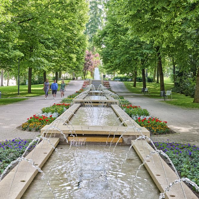 der langgezogener Brunnen mit meheren Ebenen befindet sich im Kurpark von Bad Camberg. Viele kleine Fontänen lassen stets von links und rechts neues Wasser in den Brunnen fließen. Um den Brunnen herum befinden sich links und rechts ein Weg sowie viele große Laubbäume. 