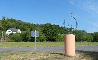 Das Foto zeigt eine Skulptur am Ortseingang von Aarbergen. Im Hintergrund ist zwischen Bäumen ein Wohnhaus zu sehen. Der Himmel ist blau. 