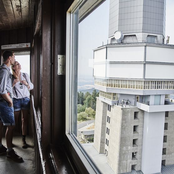 Ein Mann und eine Frau stehen im Aussichtsturm auf dem großen Feldberg und schauen nach draußen. Im Hintergrund ist ein weiterer Turm zu sehen, der zum Hessichen Rundfunk gehört. 