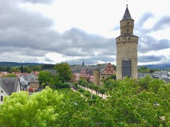 Im Vordergrund sind Baumkronen zu sehen, dahinter rechts im Bild der Hexenturm in Idstein, links davon Dächer der Altstadt. Der Himmel ist bewölkt.