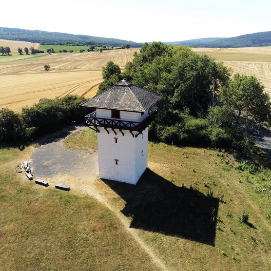 Drohnenaufnahme des Turm Dasbach. Der Turm ist quadratisch und weiß mit dunklem Dach. Außen herum ist vor allem Wiese und wenige Bäume zu sehen. 