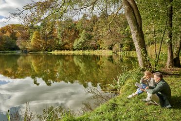 Ein Mann und eine Frau sitzen am Ufer des Hattsteinweihers. Der Wald ist in herbstliche Farben getaucht. Die Bäume rund um den See spiegeln sich im Wasser. 